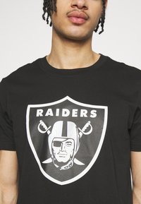 Camiseta de algodón negra con un prominente logo blanco de los Raiders en un diseño de escudo, que incluye una figura de pirata estilizada y espadas cruzadas.