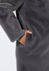 Abrigo de piel sintética gris con una textura suave, con mangas largas y un bolsillo lateral. La tela tiene un aspecto suave y aterciopelado.