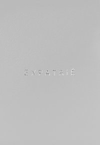 Szürke bőrtermék, puha textúrával, középen dombornyomott ezüst szöveggel "EXPATRIÉ". Minimalista dizájn és finom fényesség.