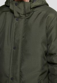 Kurtka puchowa w oliwkowej zieleni z wysokim kołnierzem, zapinana na guziki, o teksturowanej tkaninie. Posiada kaptur i gładkie, błyszczące wykończenie.