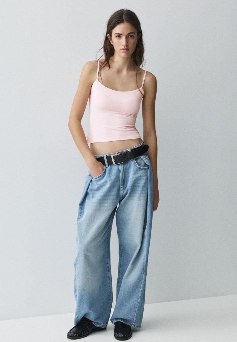 Jeune femme portant un débardeur rose clair et un jean bleu clair à jambes larges avec une ceinture noire et des chaussures noires, debout devant un fond uni.