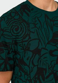 Camisa de manga corta en verde oscuro con un estampado botánico negro. Tiene un cuello redondo y una tela texturizada con un patrón sutil.