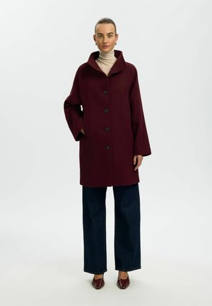 Femme portant un manteau boutonné rouge foncé sur un col roulé beige, un jean large foncé et des chaussures rouges pointues, debout devant un fond blanc uni.