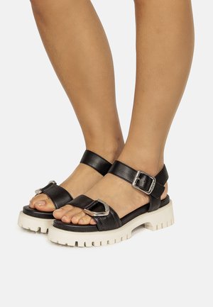 Sandales en cuir noir avec une semelle blanche épaisse, dotées d'une sangle de cheville et de boucles argentées. Design ouvert à l'avant, brides supérieures texturisées.
