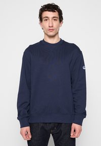 Sweatshirt bleu marine avec un col rond côtelé et des manches longues, arborant un patch logo sur la manche gauche et une finition texturée.