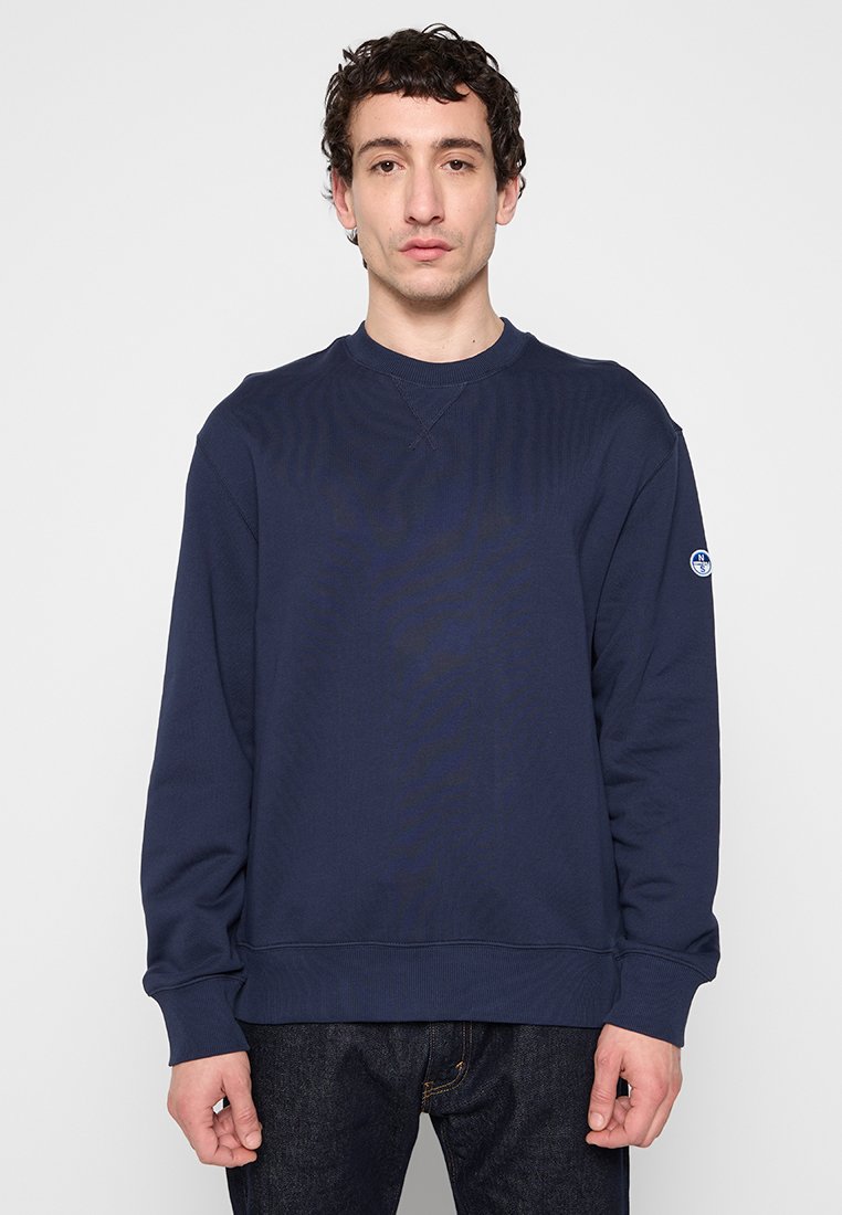 Sweatshirt bleu marine avec un col rond côtelé et des manches longues, arborant un patch logo sur la manche gauche et une finition texturée.
