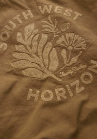 Bruine stof met beige geborduurde tekst "SOUTH WEST" en "HORIZON", met bloem- en bladmotieven. Textuur van de stiksels is goed zichtbaar.