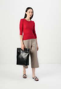 Red long-sleeve fitted top, beige cropped wide-leg trousers, holding a black leather tote bag with a cut-out handle, wearing black and white patterned slides.