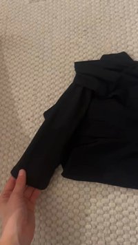 Veselă neagră cu mânecă pliată, textură netedă și fără modele vizibile. Așezată pe o suprafață texturată de culoare deschisă.