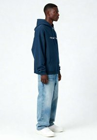 Marineblauwe hoodie met een voorzak en een trekkoord, voorzien van witte tekst. Gecombineerd met lichtblauwe jeans en witte sneakers. Zijaanzicht.