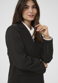 Černý blazer se strukturovanými rameny, knoflíkovými manžetami a klopami, spárovaný s bílou košilí s límečkem. Hladká tkanina a čisté linie.