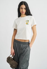 Λευκό κοντό t-shirt με ένα πράσινο και πορτοκαλί λουλουδάτο σχέδιο και λογότυπο στο στήθος, φορεμένο με φαρδιά σκούρα γκρι παντελόνια.