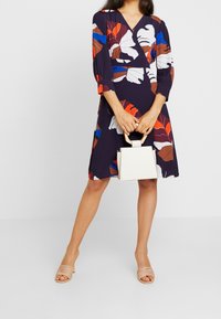 Marineblaues Kleid mit bunten Blattmustern, V-Ausschnitt, dreiviertel Ärmel, kombiniert mit einer strukturierten weißen Handtasche und beigen Sandalen mit Riemen.