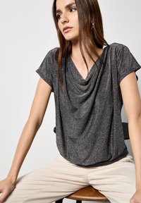 Kurzärmliges Oberteil in Dunkelgrau, mit einem drapierten Ausschnitt und weichem, schimmerndem Stoff. Getragen mit hellen, schmal geschnittenen Hosen.