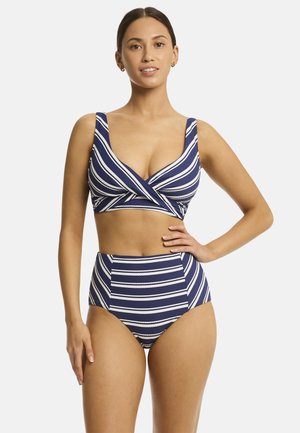 Donna che indossa un bikini a righe blu navy e bianco a vita alta, in piedi con una mano sui fianchi, su uno sfondo semplice.