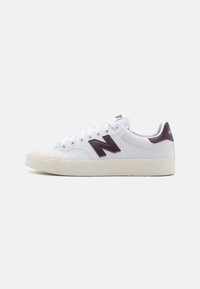 Fehér sneaker, texturált szövet felsőrésszel, sötétlila "N" logóval, gumitalppal és fehér fűzőszerkezettel. Minimalista dizájn, alacsony profil.