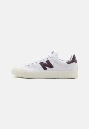 Fehér sneaker, texturált szövet felsőrésszel, sötétlila "N" logóval, gumitalppal és fehér fűzőszerkezettel. Minimalista dizájn, alacsony profil.