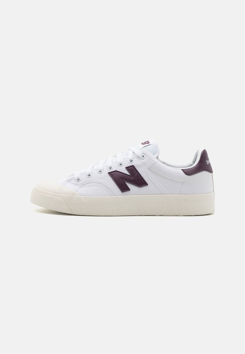 Fehér sneaker, texturált szövet felsőrésszel, sötétlila "N" logóval, gumitalppal és fehér fűzőszerkezettel. Minimalista dizájn, alacsony profil.