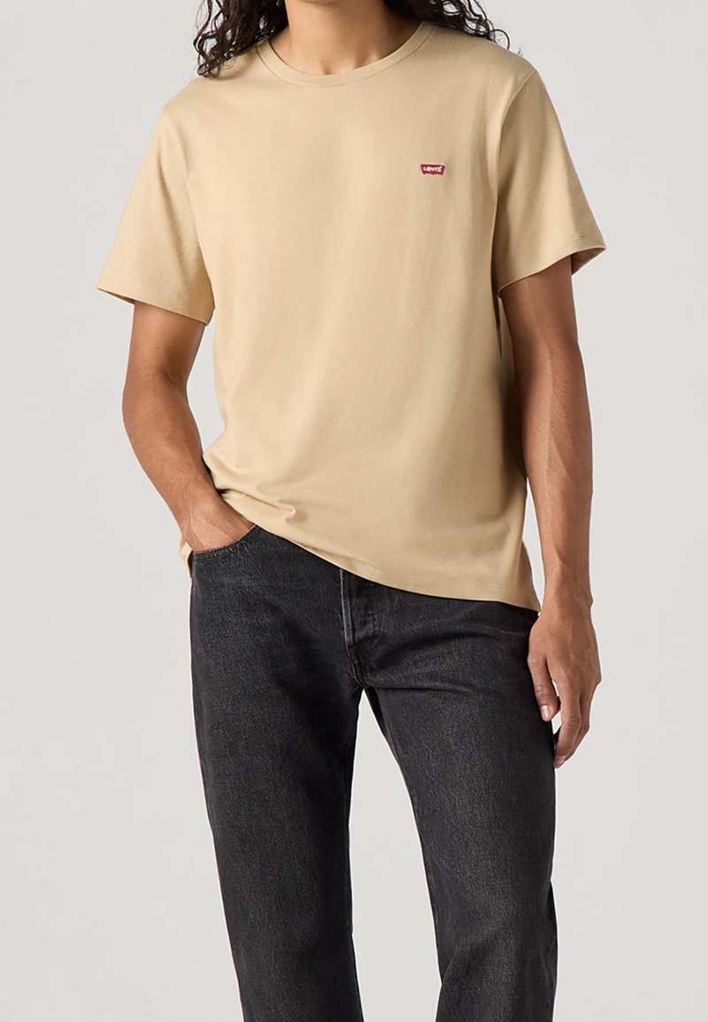 Homme portant un t-shirt beige Levi's et un jean noir, une main dans la poche, debout devant un fond uni.