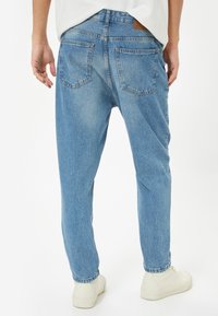 Světle modré džíny s uvolněným střihem, pěti kapsami, jemným vyblednutím a hnědou koženou nášivkou na pase. Nosené s bílými teniskami.