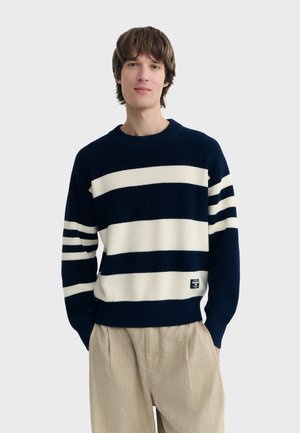 Young man wearing navy and white striped sweater and beige pants, standing with hands in pockets against plain background.