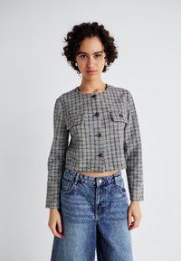 Veste cropped à carreaux gris et multicolores avec des manches longues, fermeture par bouton, et deux poches poitrine, associée à un jean bleu taille haute.