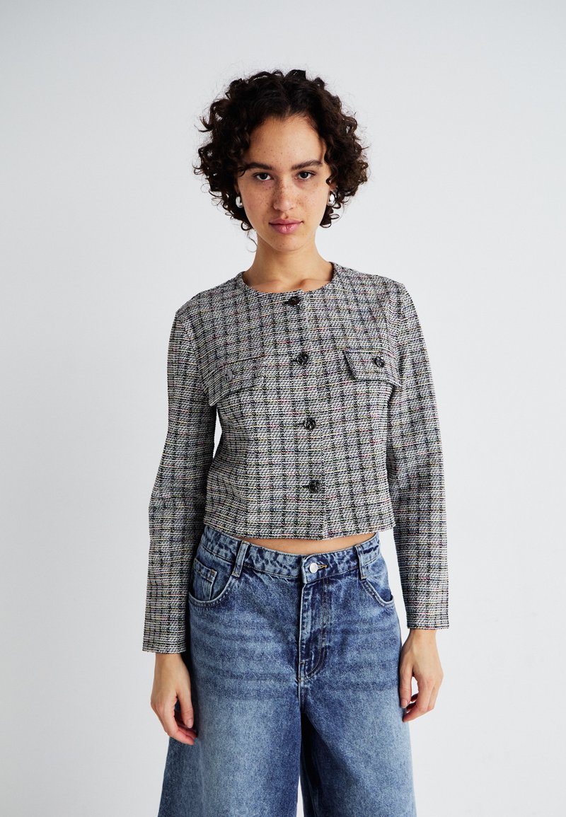 Veste cropped à carreaux gris et multicolores avec des manches longues, fermeture par bouton, et deux poches poitrine, associée à un jean bleu taille haute.
