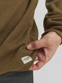 Brązowy sweter dziany z teksturowanym wzorem. Posiada metkę na dolnym brzegu z napisem "JACK & JONES EST. 1989." Ręka trzymająca materiał.