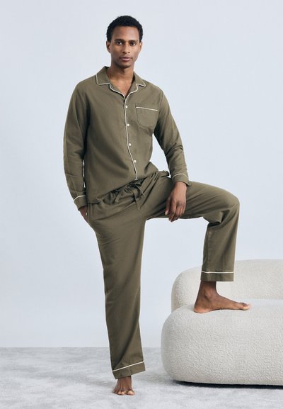 Man in olijfgroene pyjama met witte biezen, staande op blote voeten op een tapijt, met één voet op een afgeronde witte gestoffeerde stoel.