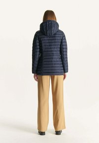 Marine pufferjack met een quilted ontwerp, een capuchon en lange mouwen, gecombineerd met tan broeken met wijde pijpen. Versterkte naden en een gladde textuur.