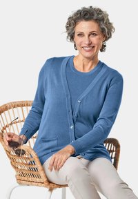 Gilet bleu clair avec fermeture à bouton et deux poches, associé à un haut assorti. Porté par-dessus un pantalon crème en étant assis dans une chaise tissée.