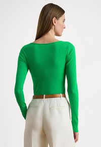 Μπλούζα με μακριά μανίκια και ριμπ ύφανση σε ζωηρό πράσινο ύφασμα με φαρδύ λαιμόκοψη, συνδυασμένη με εκρού παντελόνι και καφέ ζώνη.