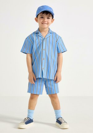 Young boy smiling, wearing blue and white vertical striped short-sleeve shirt and matching shorts, blue cap, socks, and navy sneakers.