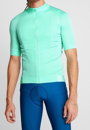 Mann trägt ein mintgrünes kurzärmeliges Radtrikot mit Frontreißverschluss und blaue gepolsterte Radhosen und steht vor einem einfarbigen Hintergrund.