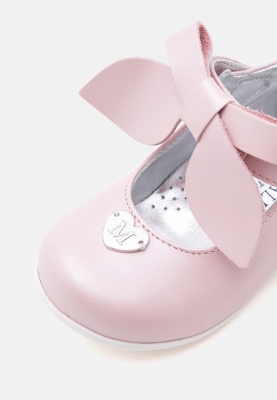 Chaussure enfant rose clair avec détail nœud et charme métallique en forme de cœur gravé de la lettre M à l’avant.