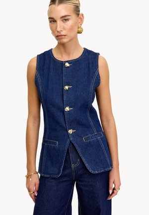 Femme portant un gilet en denim sans manches bleu foncé avec des boutons dorés et un pantalon en denim assorti, accessoirisé avec des bijoux en or.