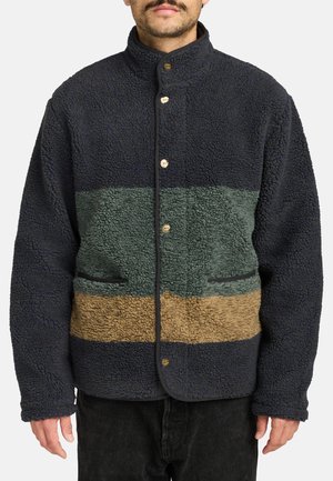 Mężczyzna ubrany w granatowo-zielono-brązową kurtkę z polaru sherpa, zapinaną na guziki, z dwoma przednimi kieszeniami, stoi na białym tle.