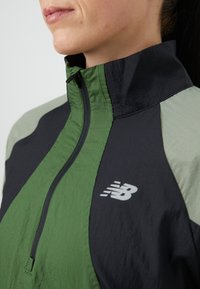 Veste à demi-zip avec un mélange de panneaux vert foncé, noir et gris clair, fabriquée en tissu léger et texturé, avec un logo sur la poitrine.