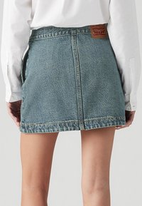Džínová sukně ve vybledlé modré barvě s vysokým pasem, roztrhaným lemem a šitými detaily. Na zadní straně se nachází hnědá kožená nášivka.