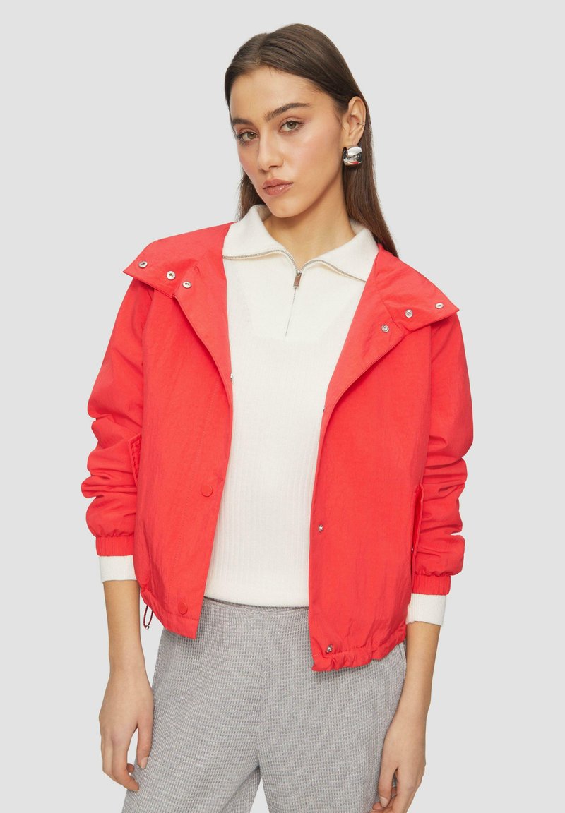 Raudona lengva striukė su užsegimais ir reguliuojamu apačiu, su gobtuvu ir elastingomis rankovėmis. Modelis vilki baltą marškinėlį ir pilkas kelnes.