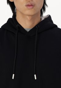 Haut à capuche noir avec un cordon de serrage, doté d'une texture douce et d'un col haut. Les cordons de serrage ont des embouts en plastique blanc.