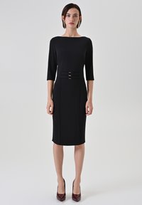 Robe moulante noire avec un ourlet arrivant au genou, des manches trois-quarts et des accents à double boucle à la taille. Tissu lisse et texturé.