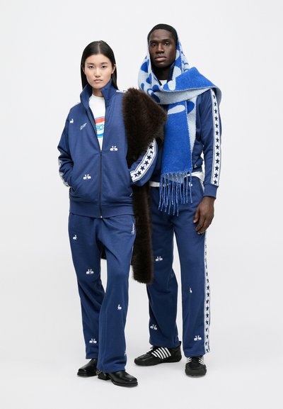 Set de îmbrăcăminte sport albastru cu motive brodate albe. Jacheta are fermoar și guler în picioare; pantalonii au dungi laterale cu model de stele. Accesoriile includ o eșarfă albastră.