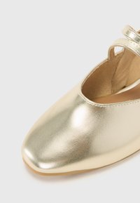 Chaussures métalliques dorées avec un bout arrondi, une texture lisse, et une sangle à la cheville. L'intérieur présente une doublure douce beige.