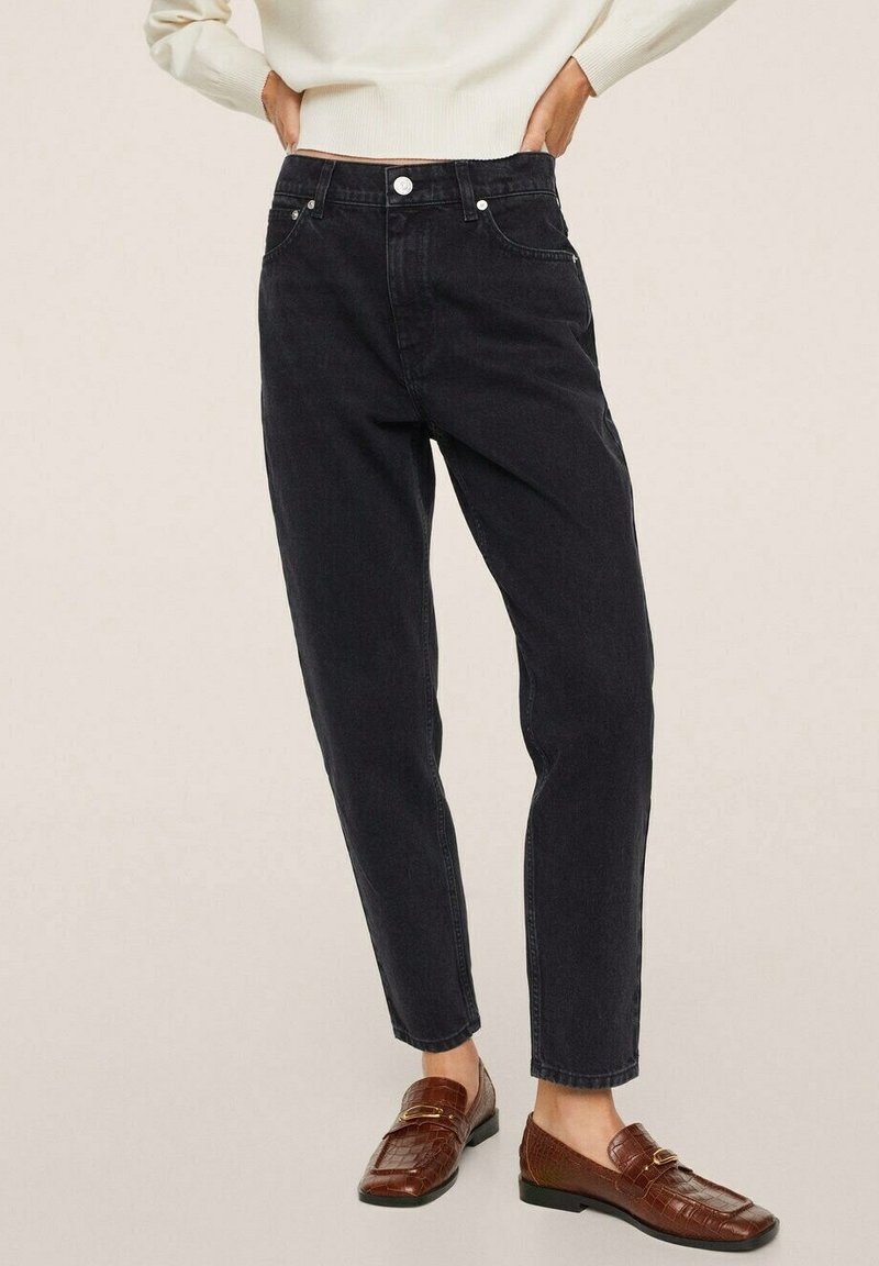 Mains sur les hanches portant un jean noir taille haute, chevilles courtes, mocassins en cuir marron et un haut à manches longues crème sur un fond beige.