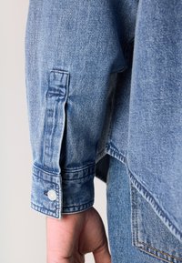 Lichtblauwe denim jacksleeve met een knoopsgatmanchet, met zichtbare stiksels en een licht vervaagde textuur.