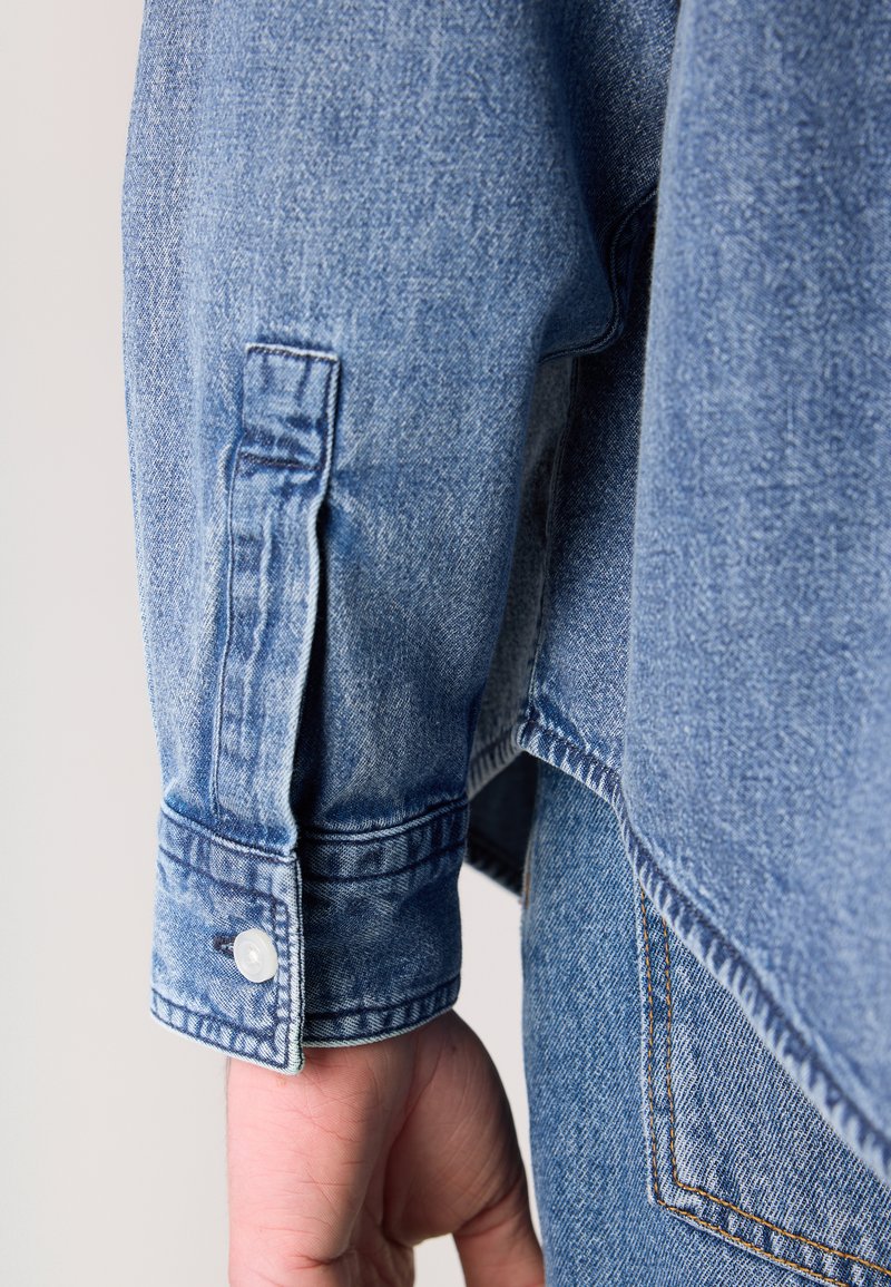 Himmelblaues Jeansjackenärmeln mit einem geknöpften Bündchen, das sichtbare Nähte und eine leicht ausgebleichte Textur aufweist.