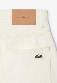 Jean en denim blanc avec poche arrière, logo crocodile vert Lacoste et patch en cuir beige cousu avec le nom de la marque au-dessus de la ceinture.