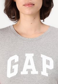 Tricou gri din bumbac, cu litere mari albe „GAP” pe piept. Guler rotund și mâneci scurte. Textură moale.