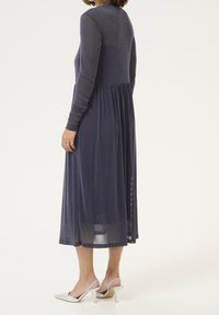 Marinblå långärmad klänning med figurnära överdel och flytande, rynkad kjol. Glänsande tyg och längd till mitt på vaden. Matchad med silverfärgade slingback-klackar.